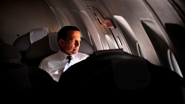 Tony Abbott appears to look out the closed window of a private jet whilst shining a light into his own face
